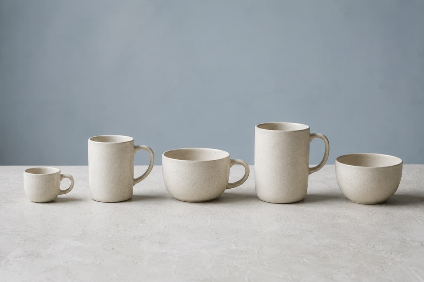 Five stoneware cups of different shapes and sizes arranged in a row on a pale worktop