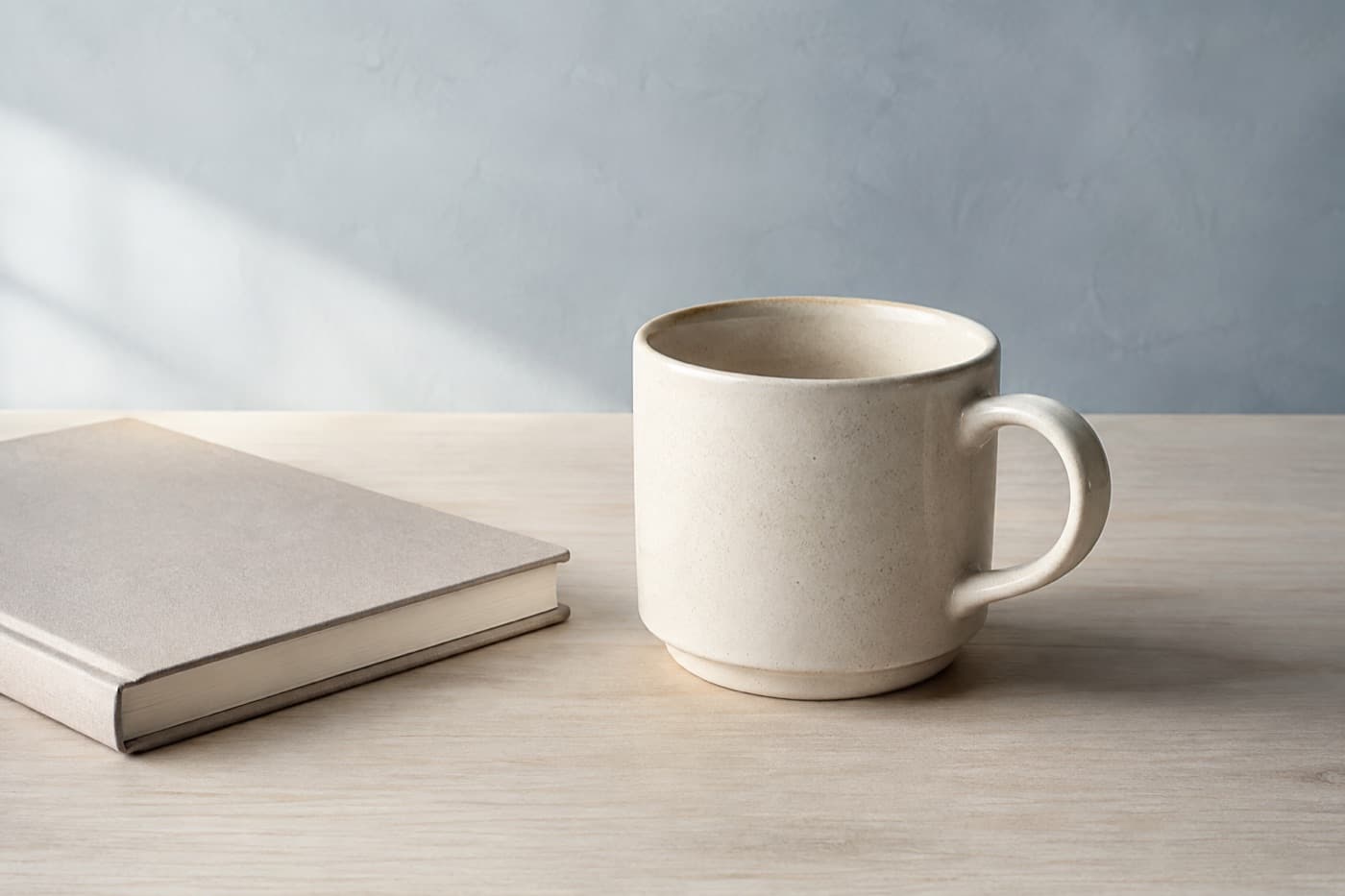 A single stoneware mug on a pale wooden desk beside a closed notebook in morning light